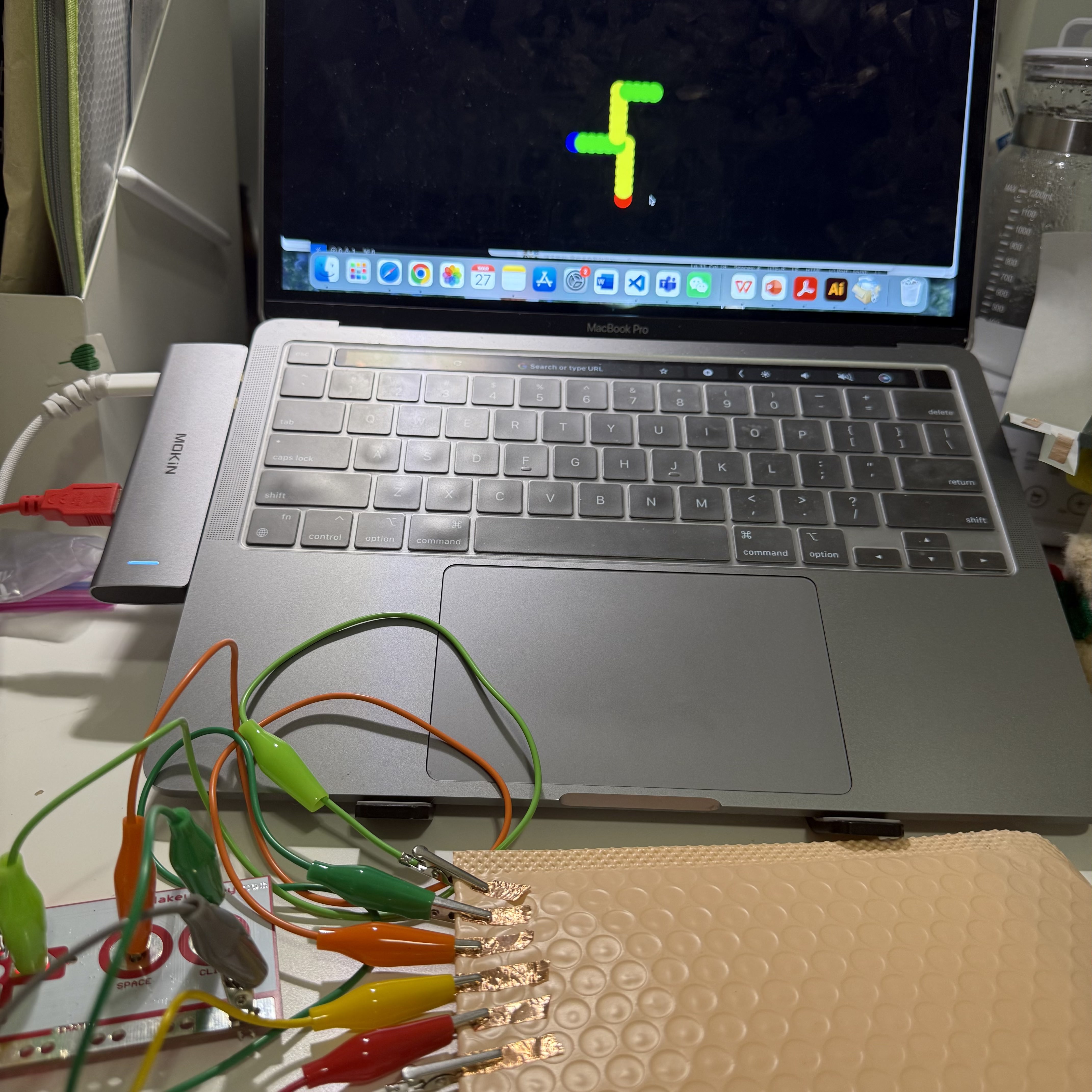In my setup, I have a Makey Makey board connected to a laptop, showing a colorful snake game on the screen. The board is hooked up to several pieces of copper tape on a textured surface, which are used as controls for the game. When I touch these copper strips, it causes the snake on the screen to move.